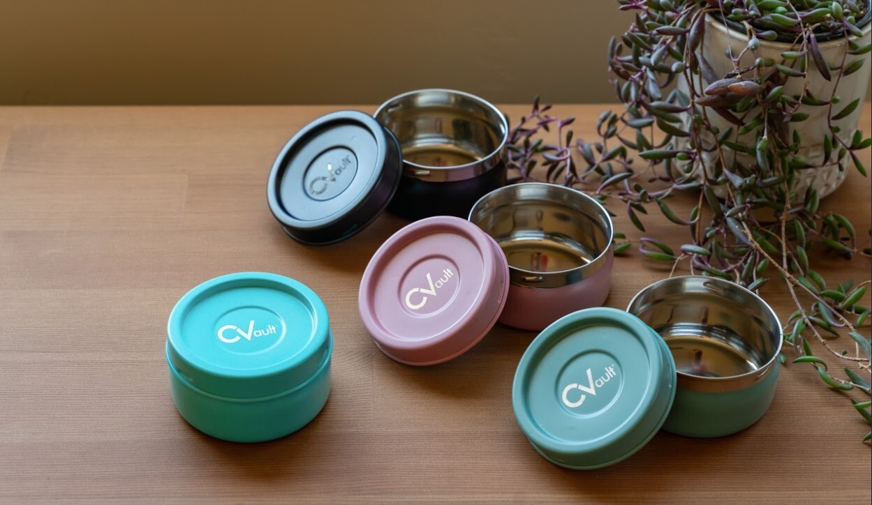 Four CVault Chromatic Collection Storage Containers sit on a wooden surface. There are four CVault colors: green, pink, black, and blue.