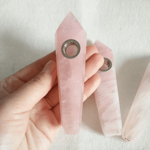 A hand holding a rose quartz pipe with a prominent point on the bowl end. There are two similar pipes on white background below.