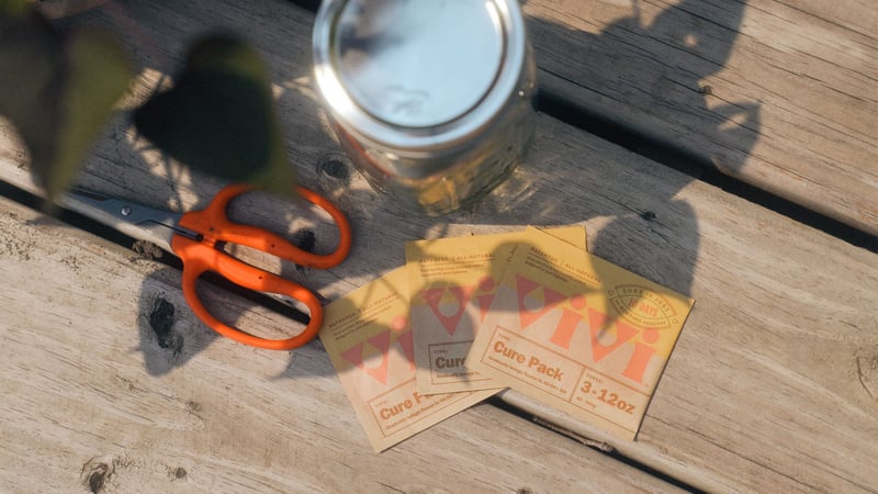 Three Vivi Cure pack laid out on a wooden surface in the sun next to a glass jar and orange trimming shears.